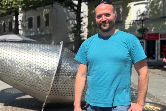 Christian Hüther steht auf dem Postplatz in Schwerte. Er trägt ein blaues T-Shirt und Jeans. Hinter ihm ist die Brunnenskulptur zu sehen.