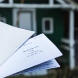 Im Vordergrund sind ein Briefumschlag und ein Bescheid über die Grundsteuer für das Jahr 2025 zu sehen, datiert vom 25.10.2024. Im Hintergrund befindet sich ein grünes Gebäude mit weißen Akzenten.