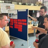 Zwei Bergkamener Bürger und Mitarbeiter des Rathauses stehen vor einer Stellwand und diskutieren Ideen, die auf roten Zetteln notiert wurden.