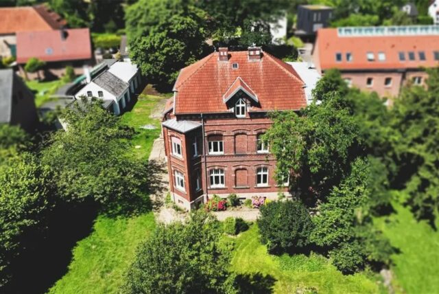 Der Hof Neuhoff in Dortmund-Eichlinghofen: Zu sehen ist ein dreistöckiges Backsteingebäude mit braunem Ziegeldach. Ein grüner Rasen umgibt das Gebäude, das über einen überdachten Eingangsbereich verfügt.
