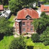 Der Hof Neuhoff in Dortmund-Eichlinghofen: Zu sehen ist ein dreistöckiges Backsteingebäude mit braunem Ziegeldach. Ein grüner Rasen umgibt das Gebäude, das über einen überdachten Eingangsbereich verfügt.