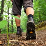 Die Beine eines Wanderes sind auf einem Waldweg zu sehen. Der Mann macht einen Schritt nach vorn und drückt sich gerade mit dem rechten Fuß ab.