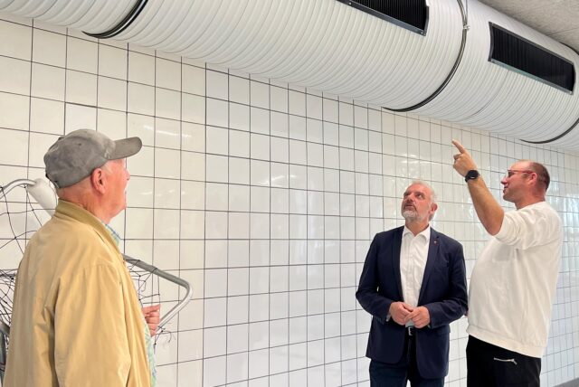 Drei Personen stehen in einem gefliesten Raum und betrachten ein Rohrsystem an der Decke. Eine der Personen deutet mit dem Finger auf das System.