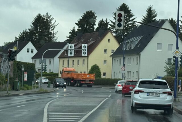 Eine nasse Straße in einer Wohngegend ist zu sehen. Mehrere Autos fahren auf der Straße, darunter ein weißer Skoda. Die Ampelanlage an der Straße ist ausgefallen.