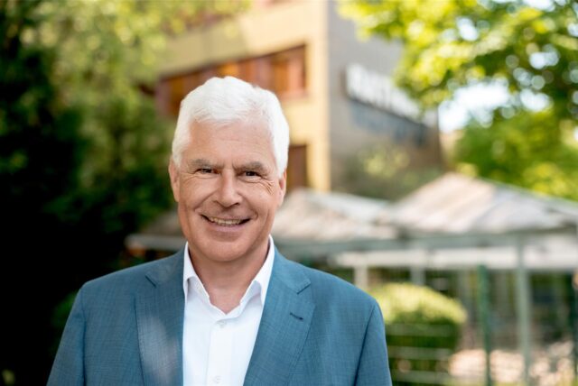 Das Bild zeigt Thomas Heinzel, der ein hellblaues Jackett und ein weißes Hemd trägt. Er lächelt in die Kamera vor einem Hintergrund aus Bäumen und einem Gebäude.