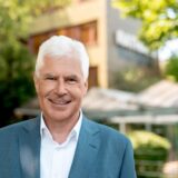 Das Bild zeigt Thomas Heinzel, der ein hellblaues Jackett und ein weißes Hemd trägt. Er lächelt in die Kamera vor einem Hintergrund aus Bäumen und einem Gebäude.