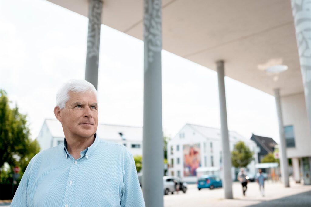 Ein Mann mit weißen Haaren und einem hellblau gestreiften Hemd steht vor einem Gebäude mit Säulen. Im Hintergrund sind weitere Gebäude, geparkte Autos und Passanten zu sehen. Ein Mann mit weißen Haaren und einem hellblau gestreiften Hemd steht vor einem Gebäude mit Säulen. Im Hintergrund sind weitere Gebäude, geparkte Autos und Passanten zu sehen.