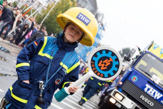 Ein junger Teilnehmer der THW-Jugend trägt eine Schutzkleidung und einen Helm mit dem THW-Logo. Er hält ein Schild mit der Aufschrift „Jugend“ und dem THW-Symbol in der Hand, während im Hintergrund ein THW-Fahrzeug zu sehen ist.