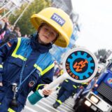 Ein junger Teilnehmer der THW-Jugend trägt eine Schutzkleidung und einen Helm mit dem THW-Logo. Er hält ein Schild mit der Aufschrift „Jugend“ und dem THW-Symbol in der Hand, während im Hintergrund ein THW-Fahrzeug zu sehen ist.