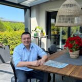 Peter Wehlack sitzt an einem Holztisch unter einer überdachten Terrasse. Auf dem Tisch liegen eine Zeitung und eine Topfpflanze mit roten Blüten.