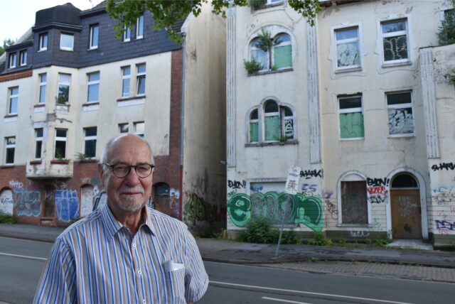 Götz Kalthoff steht an der Witttener Straße in Dortmund-Dorstfeld, im Hintergrund sind zwei leerstehende und verfallenene Häuser zu sehen.