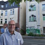Götz Kalthoff steht an der Witttener Straße in Dortmund-Dorstfeld, im Hintergrund sind zwei leerstehende und verfallenene Häuser zu sehen.