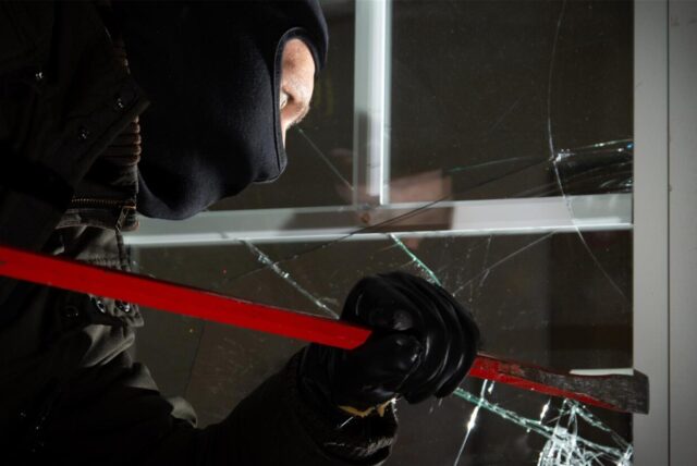 Maskierter Einbrecher hebelt mit einem Brecheisen ein Fenster auf – Glasscheibe ist bereits gesplittert.