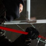 Maskierter Einbrecher hebelt mit einem Brecheisen ein Fenster auf – Glasscheibe ist bereits gesplittert.