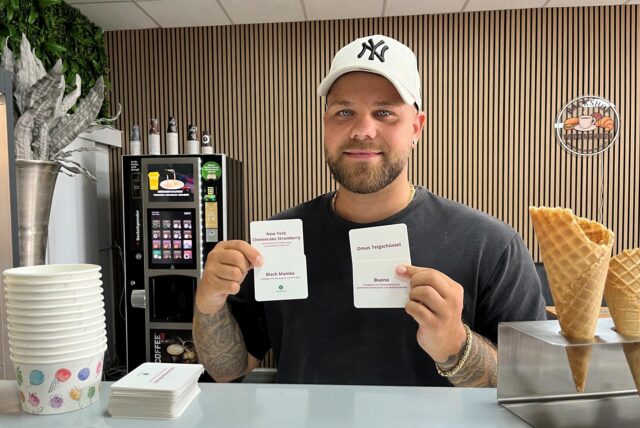 Justin Jagusch vom „Eiscafé Weddinghofen“ steht hinter dem Tresen seiner Eisdiele und hält Schilder in die Kamera, die von unterschiedlichen Geschmacksrichtungen zeugen.