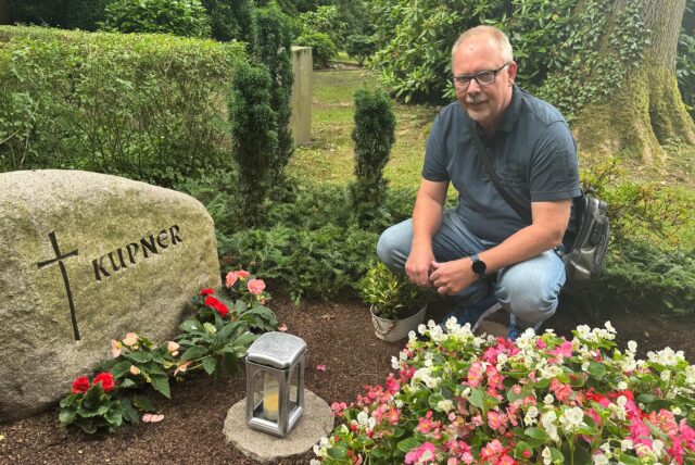 Ein Mann hockt vor einem Grabstein auf einem Friedhof. Der Grabstein trägt die Inschrift „KUPNER“ und ein Kreuzsymbol.