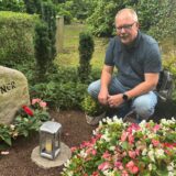 Ein Mann hockt vor einem Grabstein auf einem Friedhof. Der Grabstein trägt die Inschrift „KUPNER“ und ein Kreuzsymbol.