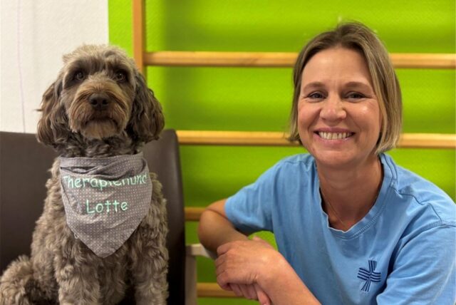 Therapiehund Lotte trägt ein Halstuch mit ihrem Namen. Neben ihr hockt Frauchen Christina Martin. Die beiden posieren vor einer grünen Wand mit Leiter im Marienkrankenhaus Schwerte.