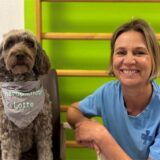 Therapiehund Lotte trägt ein Halstuch mit ihrem Namen. Neben ihr hockt Frauchen Christina Martin. Die beiden posieren vor einer grünen Wand mit Leiter im Marienkrankenhaus Schwerte.