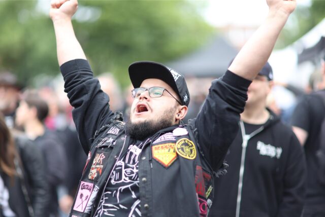 Ein Mann mit Bart, Brille und Mütze reckt vor der Alternative Stage beim Brunnenfestival in Lünen die Arme in die Höhe. Er trägt eine Jacke mit verschiedenen Aufnähern und ein schwarzes T-Shirt mit weißem Aufdruck.