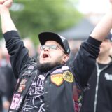 Ein Mann mit Bart, Brille und Mütze reckt vor der Alternative Stage beim Brunnenfestival in Lünen die Arme in die Höhe. Er trägt eine Jacke mit verschiedenen Aufnähern und ein schwarzes T-Shirt mit weißem Aufdruck.