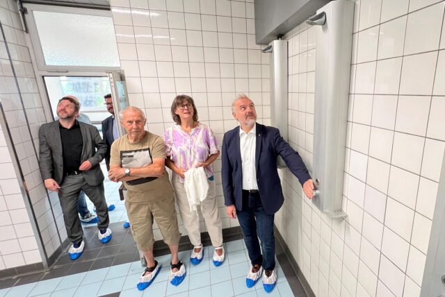 Eine Gruppe von Personen steht in einem gefliesten Raum, vermutlich im Rahmen einer Begehung oder Besichtigung. Die Personen tragen blaue Überschuhe und betrachten die sanitären Installationen an der Wand.
