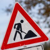Das Bild zeigt ein Verkehrsschild, das auf Bauarbeiten hinweist. Das Schild ist dreieckig mit einem roten Rand und einem weißen Hintergrund, auf dem eine stilisierte Figur mit einer Schaufel abgebildet ist.