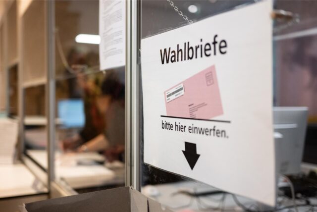 Ein Schild mit der Aufschrift „Wahlbriefe, bitte hier einwerfen“ hängt an einer Glaswand. Es zeigt eine stilisierte Darstellung eines Wahlbriefs und einen Pfeil, der nach unten weist.