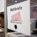 Ein Schild mit der Aufschrift „Wahlbriefe, bitte hier einwerfen“ hängt an einer Glaswand. Es zeigt eine stilisierte Darstellung eines Wahlbriefs und einen Pfeil, der nach unten weist.