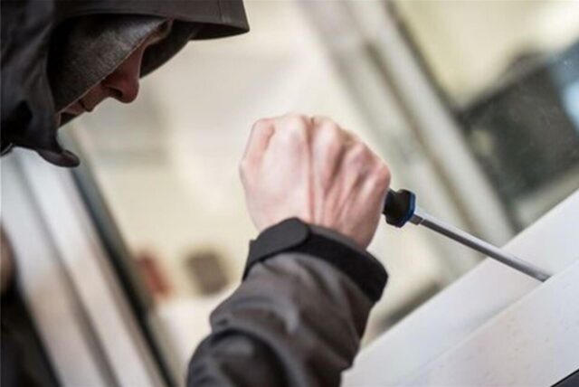 Person in Kapuzenjacke versucht mit Schraubenzieher ein Fenster aufzuhebeln – Symbolbild für Einbruch.