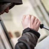 Person in Kapuzenjacke versucht mit Schraubenzieher ein Fenster aufzuhebeln – Symbolbild für Einbruch.