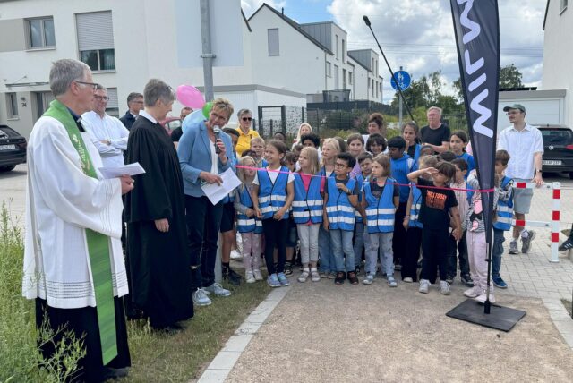 Auf einem Gruppenbild bei der Eröffnung des Spielplatzes im Wohnpark Emscherquelle in Holzwickede stehen links Pastor Bernhard Middelanis und Pfarrerin Diemut Cramer neben Bürgermeisterin Ulrike Drossel. Daneben stehen viele Kinder mit blauen Warnwesten sowie einige Betreuer und weitere Personen. Im Hintergrund sind einige weiße Häuser sowie ein bewölkter Himmel zu erkennen.
