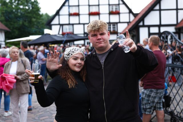 Junges Paar hebt lachend die Gläser beim Scheunenfest des Fördervereins der Freiwilligen Feuerwehr in Kamen im Freien, im Hintergrund zahlreiche Besucher vor Fachwerkhäusern in geselliger Atmosphäre.