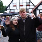 Junges Paar hebt lachend die Gläser beim Scheunenfest des Fördervereins der Freiwilligen Feuerwehr in Kamen im Freien, im Hintergrund zahlreiche Besucher vor Fachwerkhäusern in geselliger Atmosphäre.
