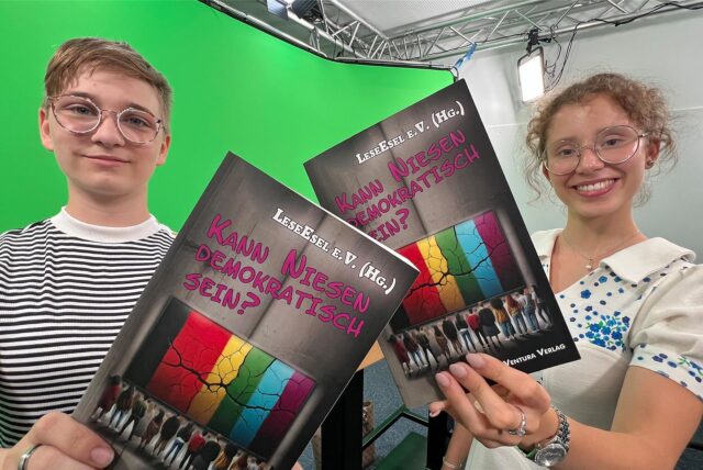 Jule Scherbes (15) und Hannah Balmaceda (16) halten vor einem Greenscreen das Demokratie-Buch in die Kamera.