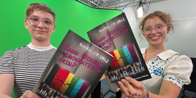 Jule Scherbes (15) und Hannah Balmaceda (16) halten vor einem Greenscreen das Demokratie-Buch in die Kamera.