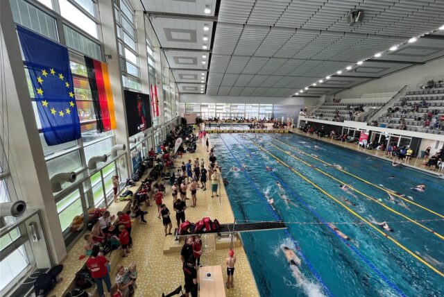 Im Südbad in Dortmund befinden sich viele Sportschwimmer im Wasser. Aktive und Trainer stehen am Beckenrand. Auf der Tribüne sitzen Zuschauer. An einer Wand hängen eine EU- und eine Deutschland-Fahne.