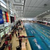 Im Südbad in Dortmund befinden sich viele Sportschwimmer im Wasser. Aktive und Trainer stehen am Beckenrand. Auf der Tribüne sitzen Zuschauer. An einer Wand hängen eine EU- und eine Deutschland-Fahne.