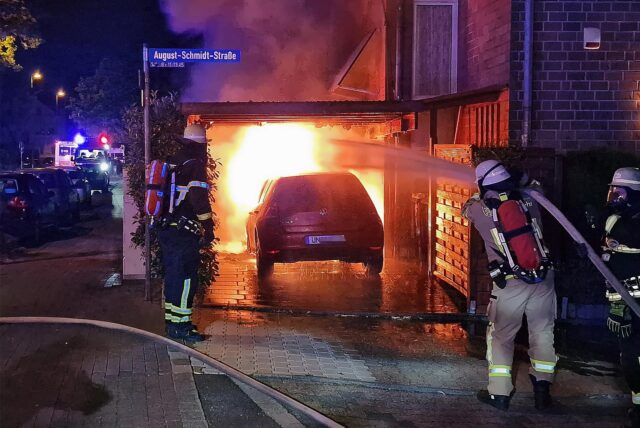 Feuerwehrkräfte in Atemschutzmontur löschen einen im Motorraum brennenden VW Golf, der unter einem Carport geparkt ist.