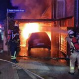 Feuerwehrkräfte in Atemschutzmontur löschen einen im Motorraum brennenden VW Golf, der unter einem Carport geparkt ist.