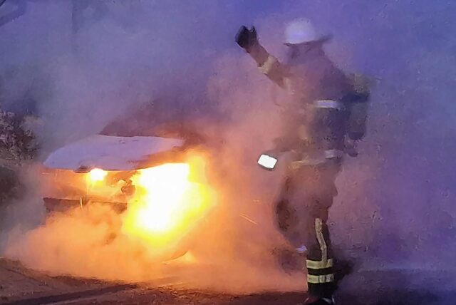 In einer dichten Rauchwolke steht ein Feuerwehrmann in Atemschutzmontur vor einem brennenden Auto.