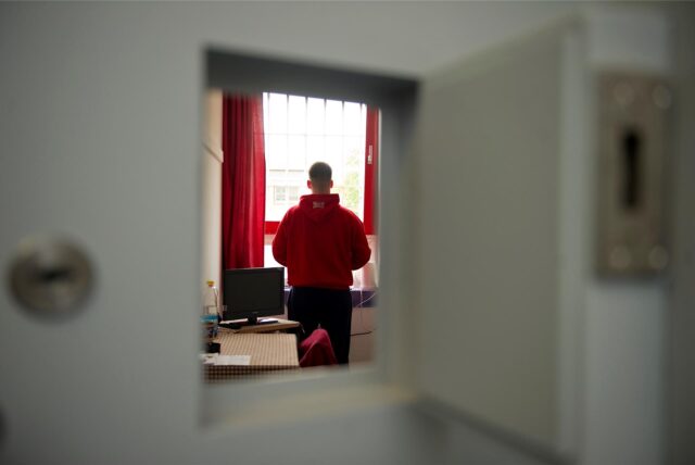 Ein Jugendlicher steht im Haftraum einer Jugendstrafanstalt mit dem Rücken zur Kamera. Er trägt einenschwarze Hose und einen roten Hoodie.