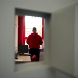 Ein Jugendlicher steht im Haftraum einer Jugendstrafanstalt mit dem Rücken zur Kamera. Er trägt einenschwarze Hose und einen roten Hoodie.