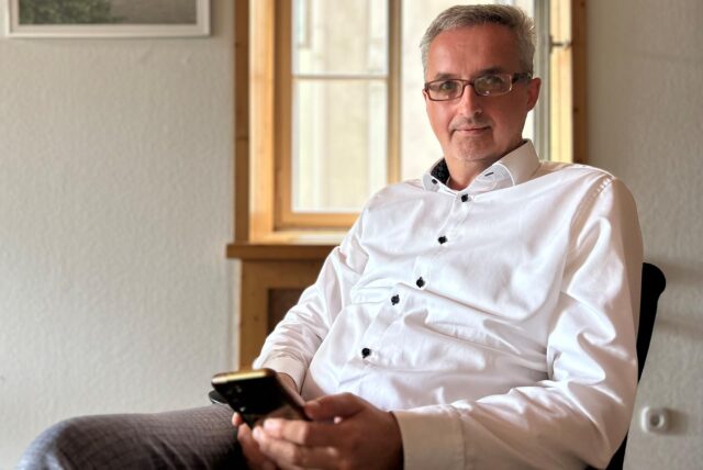 Ein Mann mit Brille sitzt in einem Stuhl. Er hält ein Smartphone in seinen Händen und trägt ein weißes Hemd.