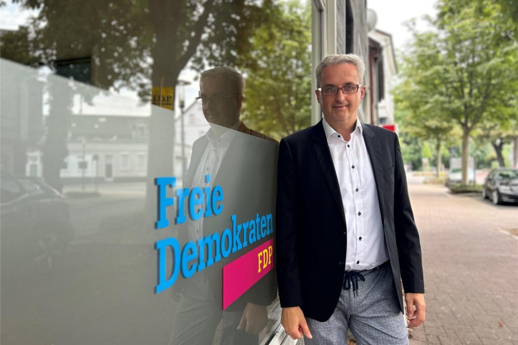 Ein Mann mit Brille und Jackett steht vor einem Gebäude. Auf dem Fenster ist der Schriftzug Ein Mann mit Brille und Jackett steht vor einem Gebäude. Auf dem Fenster ist der Schriftzug