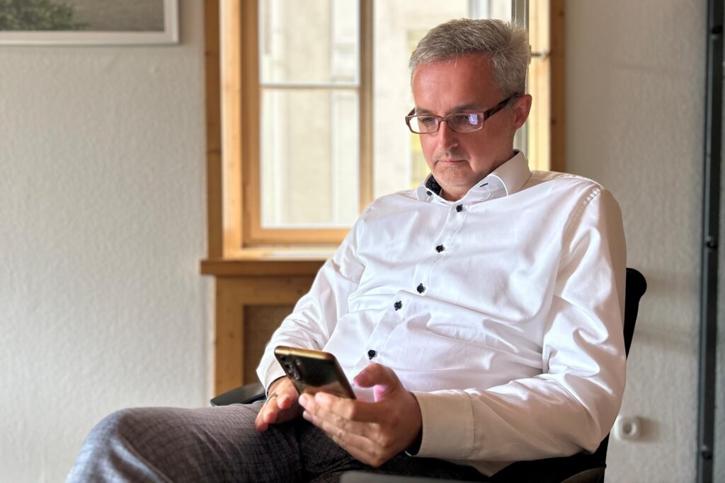Ein Mann mit Brille und weißem Hemd sitzt auf einem Stuhl und blickt auf sein Mobiltelefon. Im Hintergrund sind ein Fenster und ein gerahmtes Bild an der Wand zu sehen. Ein Mann mit Brille und weißem Hemd sitzt auf einem Stuhl und blickt auf sein Mobiltelefon. Im Hintergrund sind ein Fenster und ein gerahmtes Bild an der Wand zu sehen.