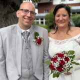 Ein Paar steht vor einem Hintergrund aus Bäumen und einem Gebäude. Die Braut trägt ein weißes Kleid und hält einen Strauß roter Rosen und Schleierkraut, während der Bräutigam einen grauen Anzug mit einer Rose am Revers trägt.