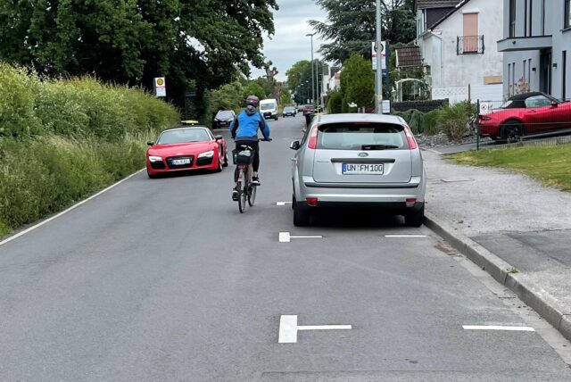 Ein Radfahrer fährt auf dem Afferder Weg zwischen einem stehenden und einem fahren Auto hindurch.