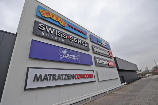 Fassadenansicht eines Gewerbe-Komplexes mit Logos mehrerer Einzelhandelsgeschäfte in Kamen am Zollpost, darunter Smyths Toys, Swiss Sense, Matratzen Concord und weitere.