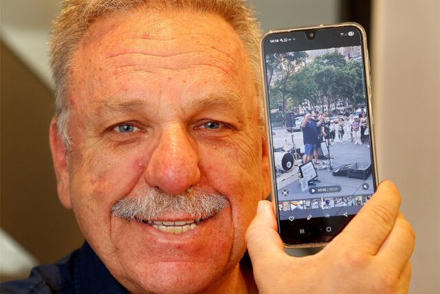 Achim Bauer mit einem Smartphone, das ihn zeigt, als er das Publikum einer Straßenband auf dem Broadway in New York anheizt.